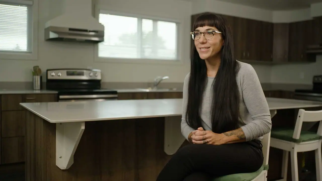 A woman sits at a bright kitchen table in a new apartment, smiling