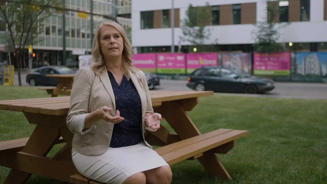 A woman speaks to camera at a picnic table in an urban park