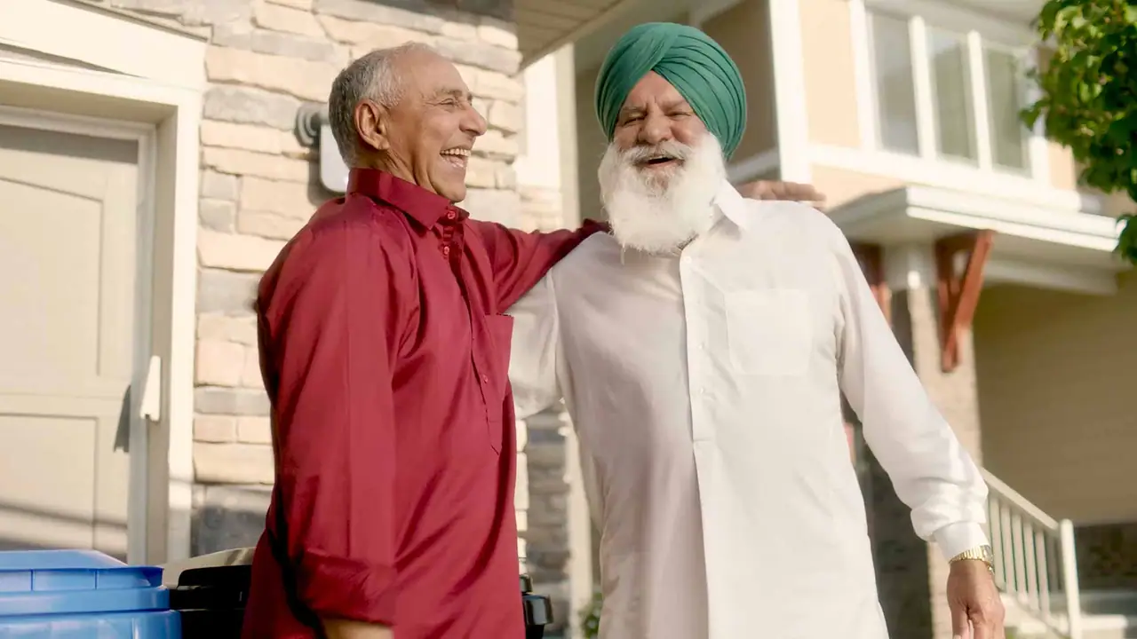 Two older men in a driveway, one in a red shirt and one in a white turban, laughing together